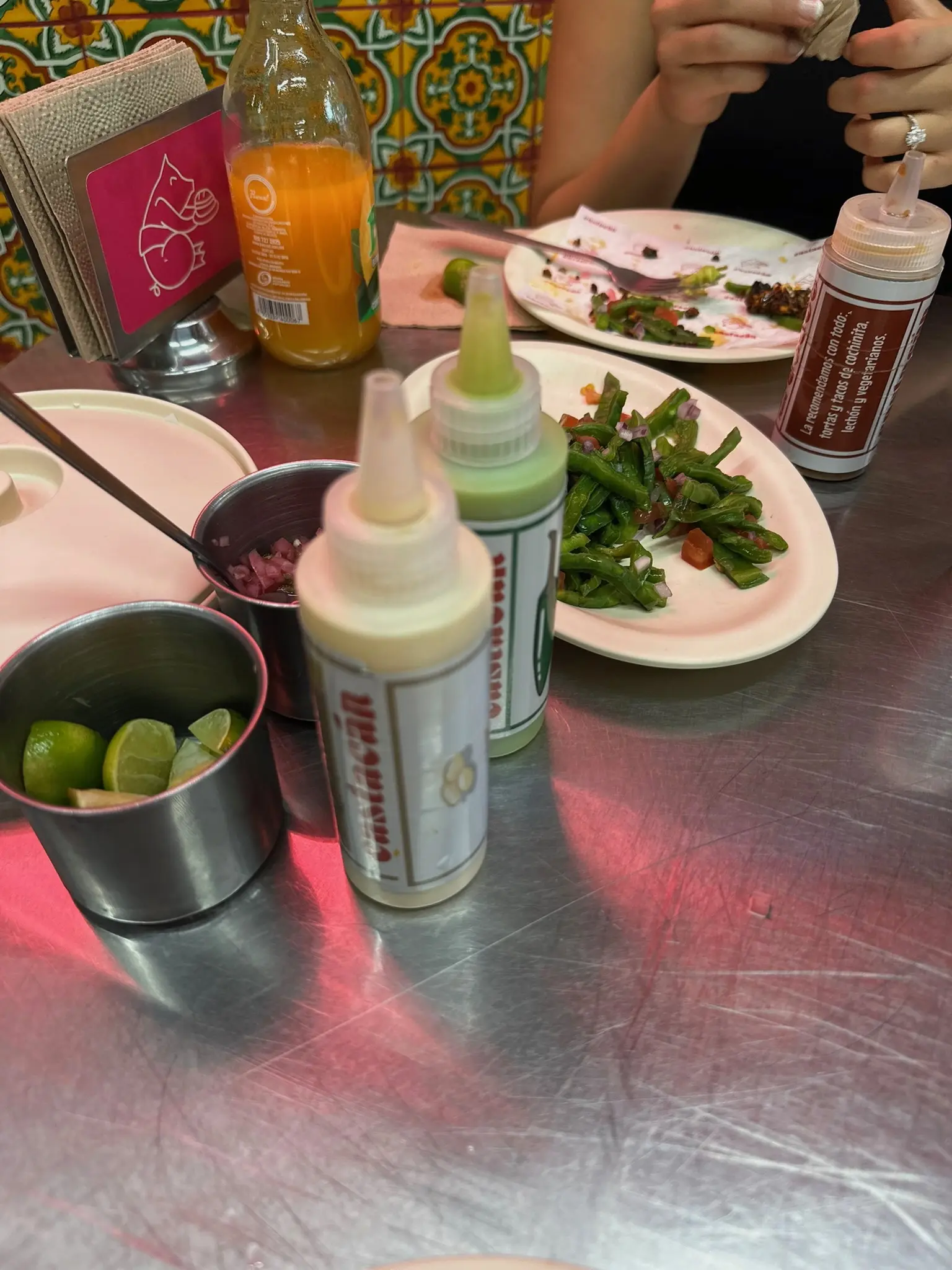 A silver metal table with a white and green salsa, a plate of nopales, a cup of limes, a cup of dices red onions, some empty plates, an orange juice, and a napkin holder with a pink image of the same pig.