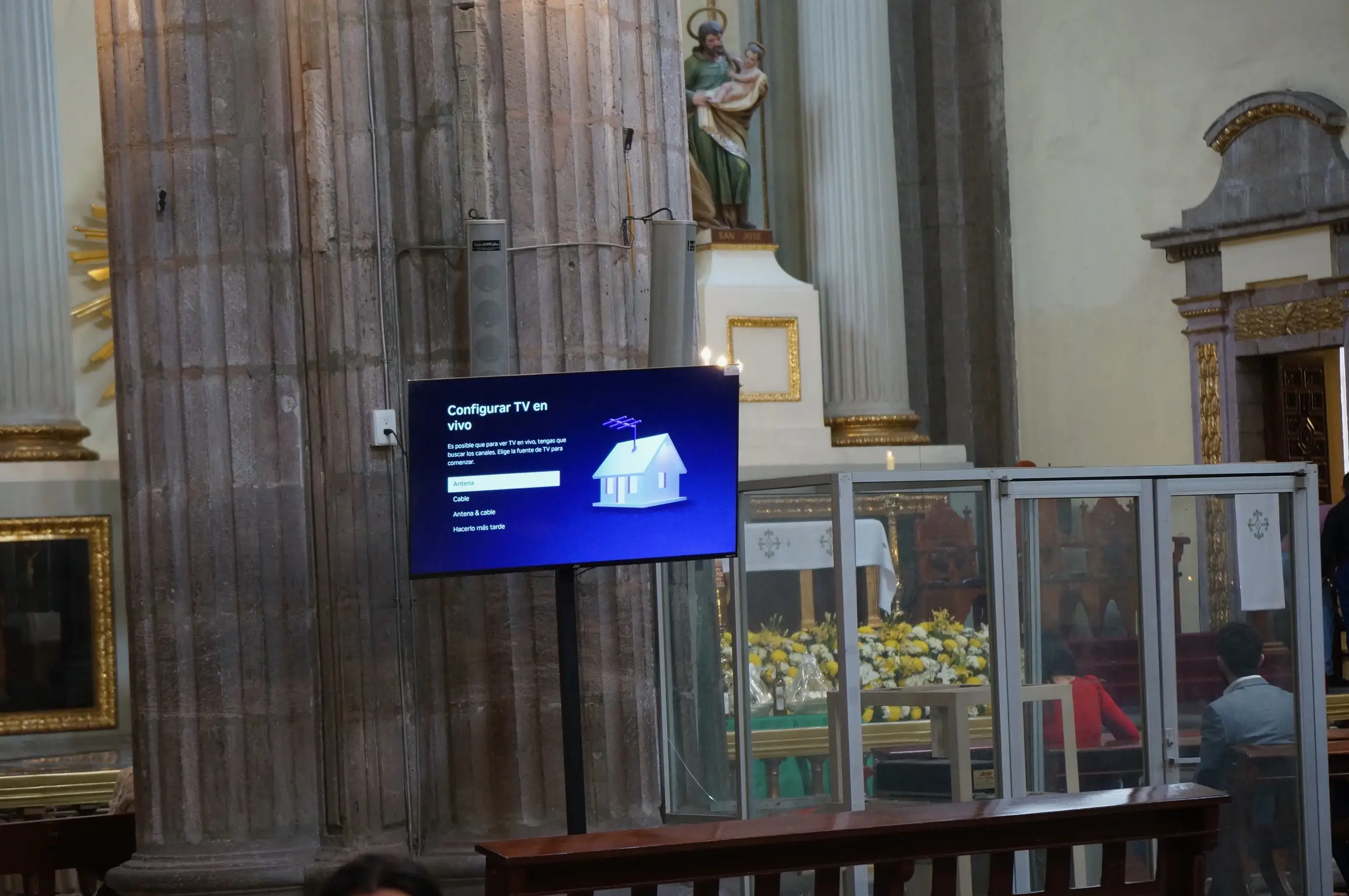 In the foreground, a TV with a purple screen says "Configurar TV en vivo: Es posible que para ver TV en vivo, tengas que buscar los canales. Elige la fuente de TV para comenzar. - Antena - Cable - Antena & cable - Hacerio más tarde". Behind the TV is a white and gold statue of Jesus with other ornate and gold-plated decorations beside it.