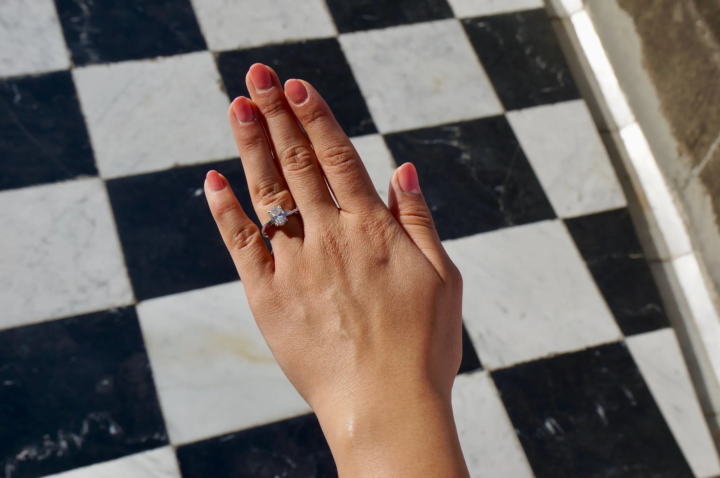 A woman's hand with pink-painted nails hovers above a black and white checkered floor. On her ring finger is a diamond engagement ring.