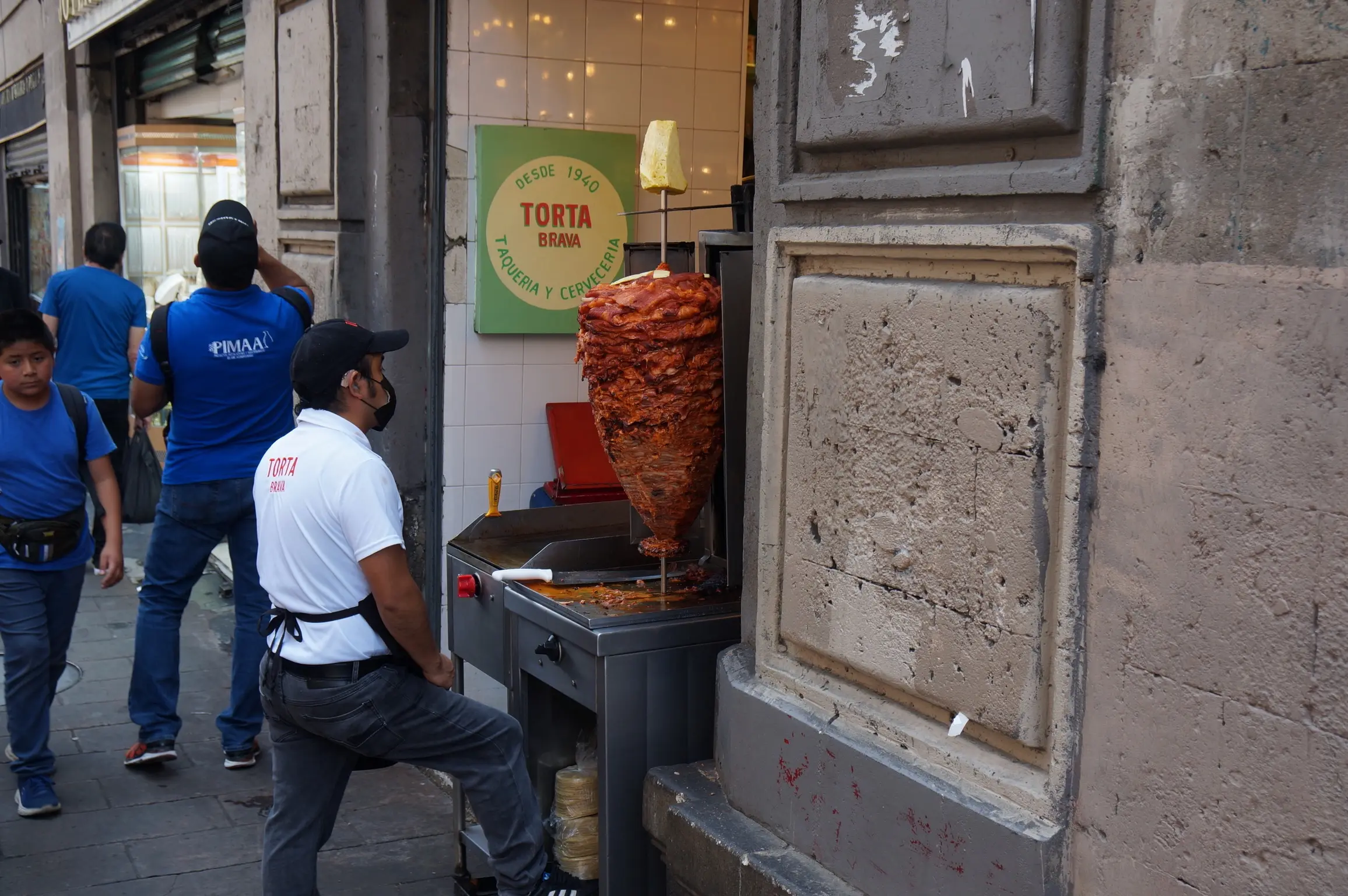 A man in a white shirt stands on the street next to a tropo of red meat, large at the top and thinner at the bottom, forming a cone shape. The sign on the side of the wall reads "Desde 1940. Torta Brava. Taqueria y Cerveceria. Several people in blue shirts walk on the sidewalk behind him."