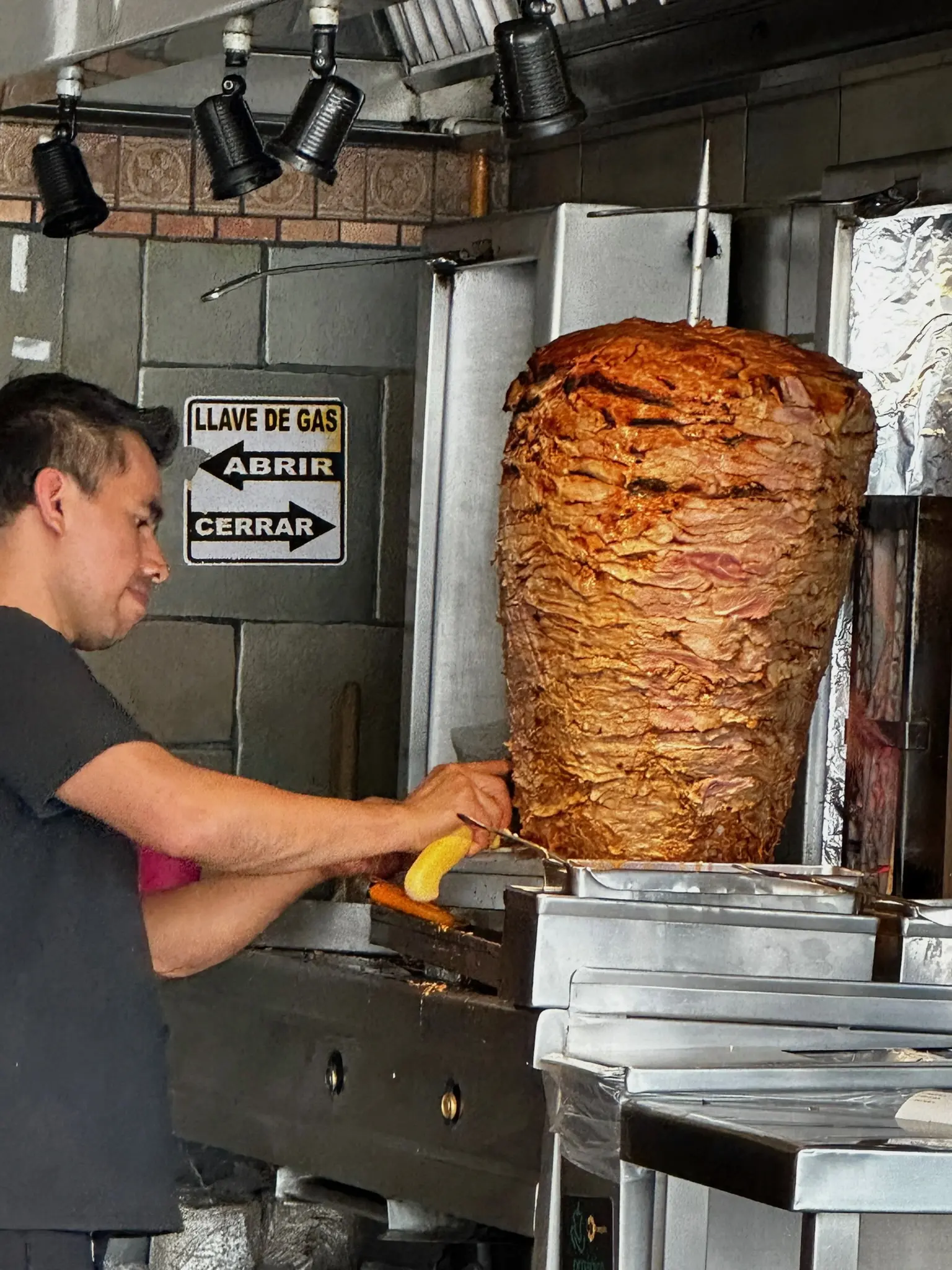 A man with short black hair in a black shirt shaves meat off of a tropo with a cone of red meat on it.