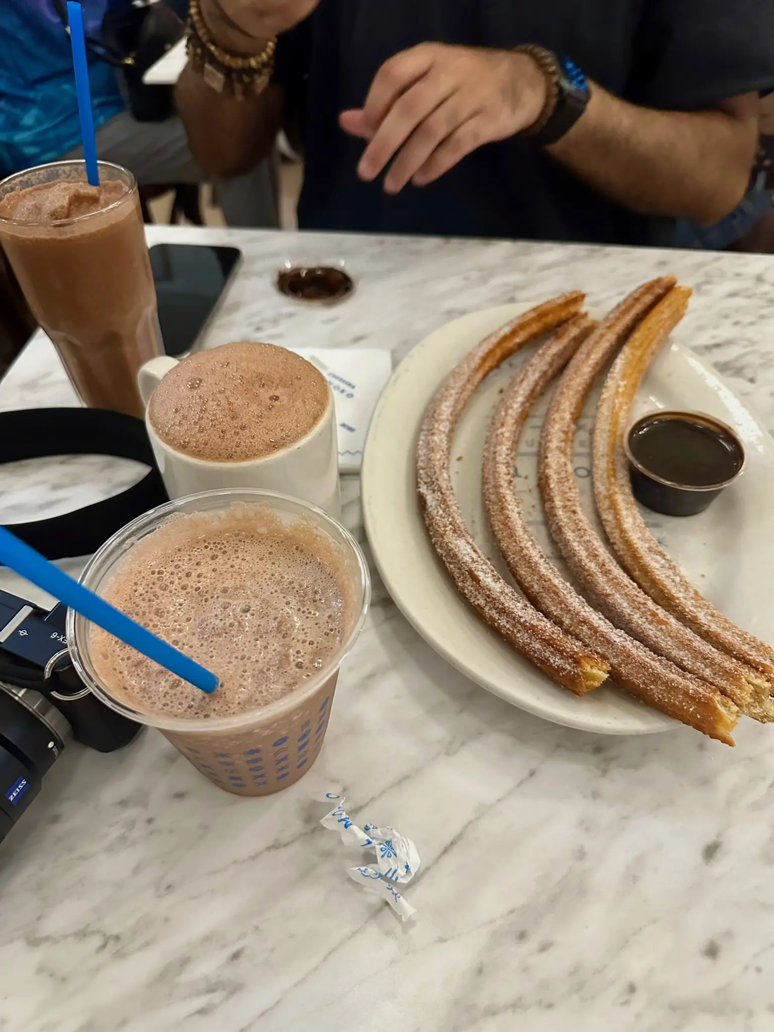 On the white marble table in front of him is a phone, a chocolate frappachino with a blue straw in a glass, a hot chocolate in a white mug, a white plate with 4 churros and a cup of dark brown chocolate sauce on it, a plastic cup with a blue straw and a chocolate milkshake in it, and a black mirrorless Sony NEX-6 camera.