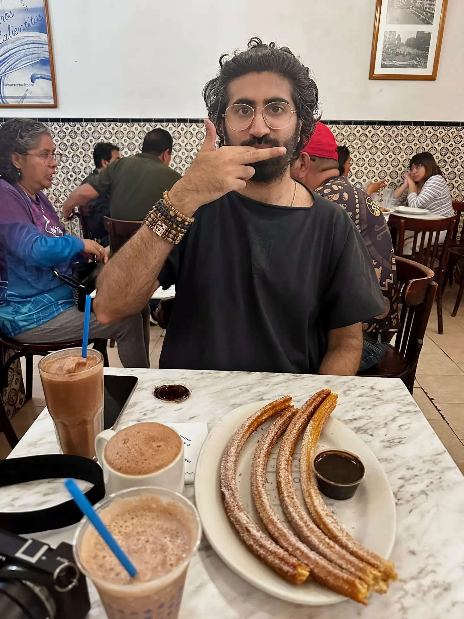 A man with long black hair put up and a black beard in a black shirt is giving the middle finger to the camera over his mouth. His wrist has several bracelets on it. On the white marble table in front of him is a phone, a chocolate frappachino with a blue straw in a glass, a hot chocolate in a white mug, a white plate with 4 churros and a cup of dark brown chocolate sauce on it, a plastic cup with a blue straw and a chocolate milkshake in it, and a black mirrorless Sony NEX-6 camera.