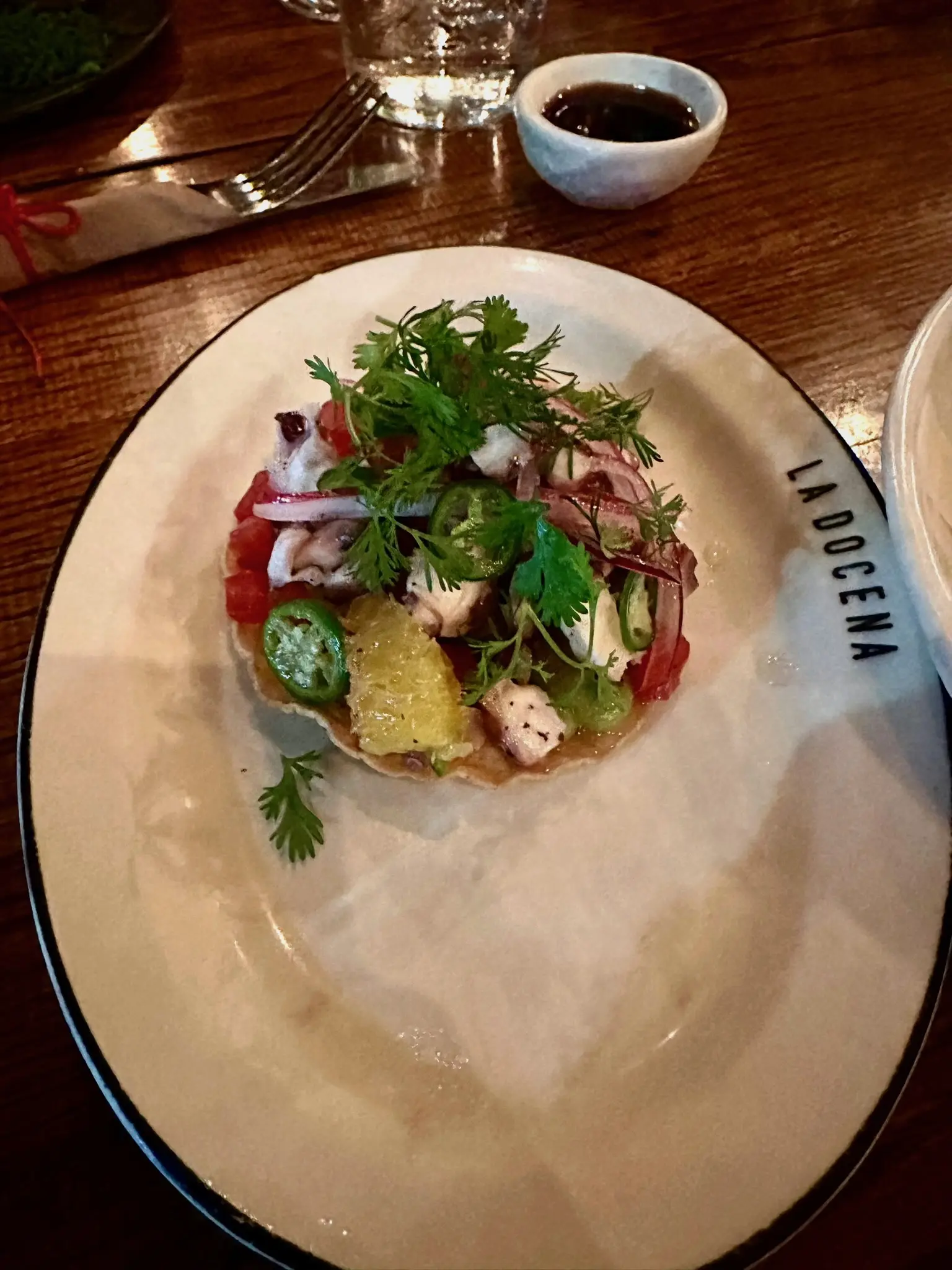 An ovular white plate with the text "LA DOCENA" on the rim sits under a tostada eith pineapple, red onion slices, jalapeño slices, grilled octopus slices, and topped with cilantro.