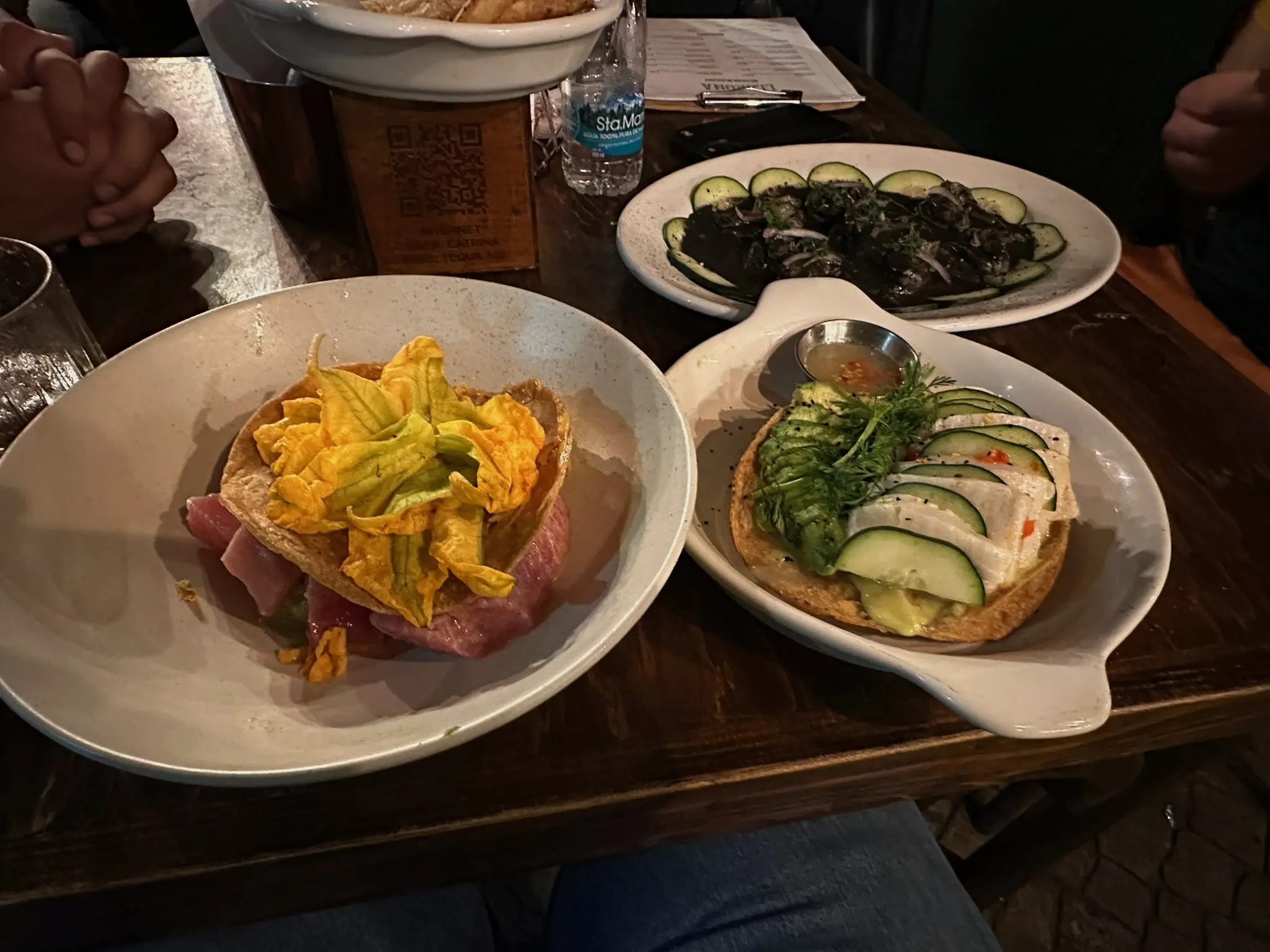 A tuna tostada with squash blossoms on top in a white bowl next to a white fish tostada on a white plate with a lip on either side of it. In the background is a white ovular plate with shrimp aguachiles negros.