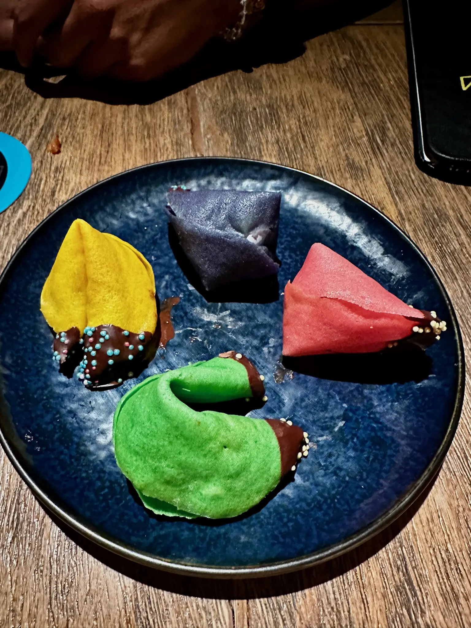 On a round blue plate sits a yellow, green, pink, and dark blue fortune cookie, each with the tip dipped in chocolate and blue sprinkles.