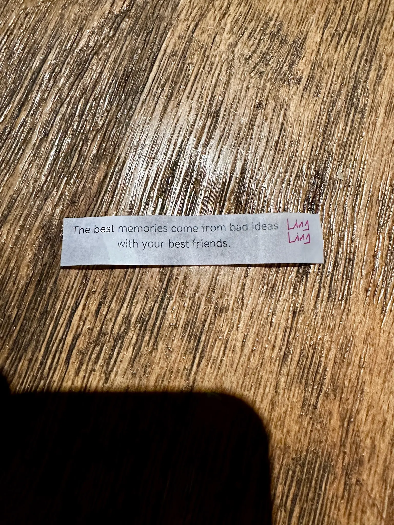 A strip of paper sits on a wooden table. The paper says "The best memories come from bad ideas with your best friends." with the Ling Ling logo in the upper right corner.