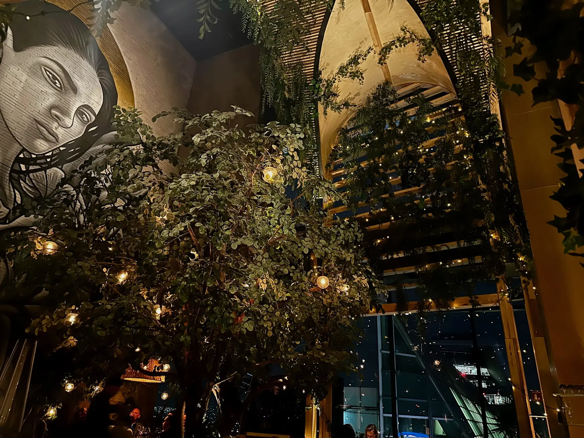 A mural of a woman with long black hair is on a wall with a large lush tree in the center of the room. The window on the right of the room has a large bamboo arch over it with bamboo slits in it.