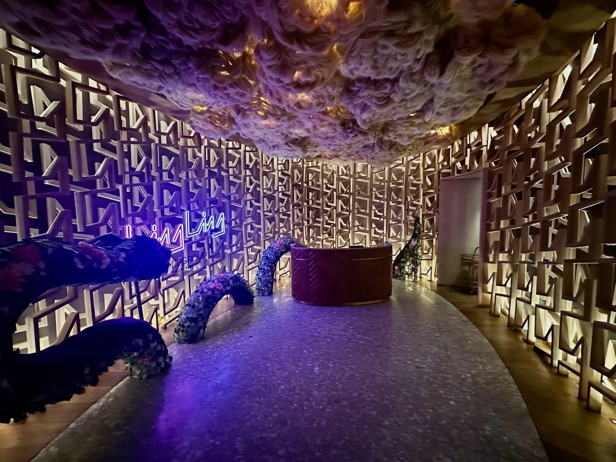 A dimly lit lobby with a fake glowing cloud ceiling and a patterned, textured wall with a blue neon sign that says "Ling Ling" on the wall on the left. Around the sign is a dragon made out of a topiary that looks like it's dissapearing into and out of the floor.