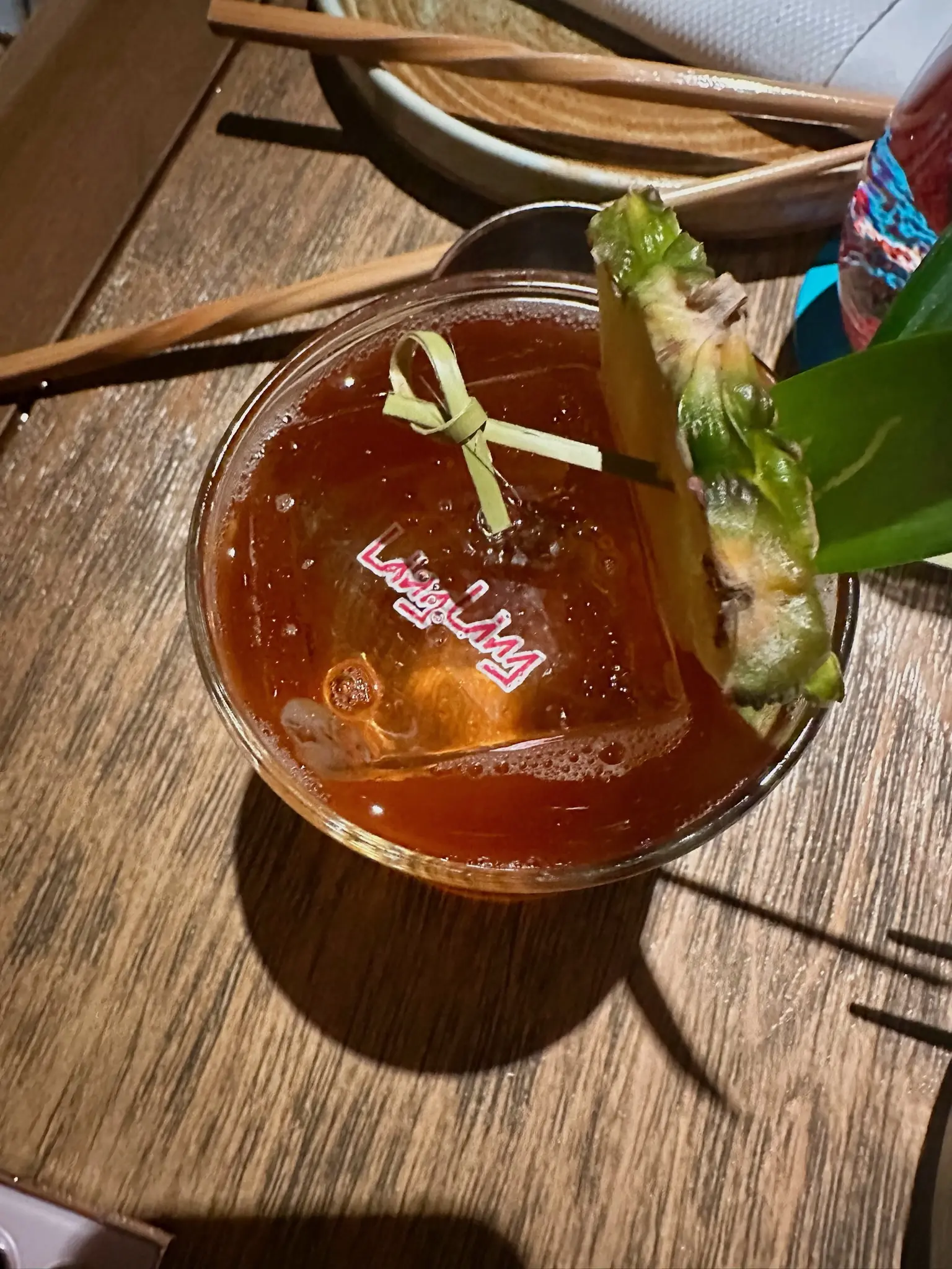 Top down on a redish brown drink with a pineapple slice, agave leaf, and bamboo toothpick in it. A large singular ice cube is in the center of the glass an the words "Ling Ling" appears in the center in pink with a white border.