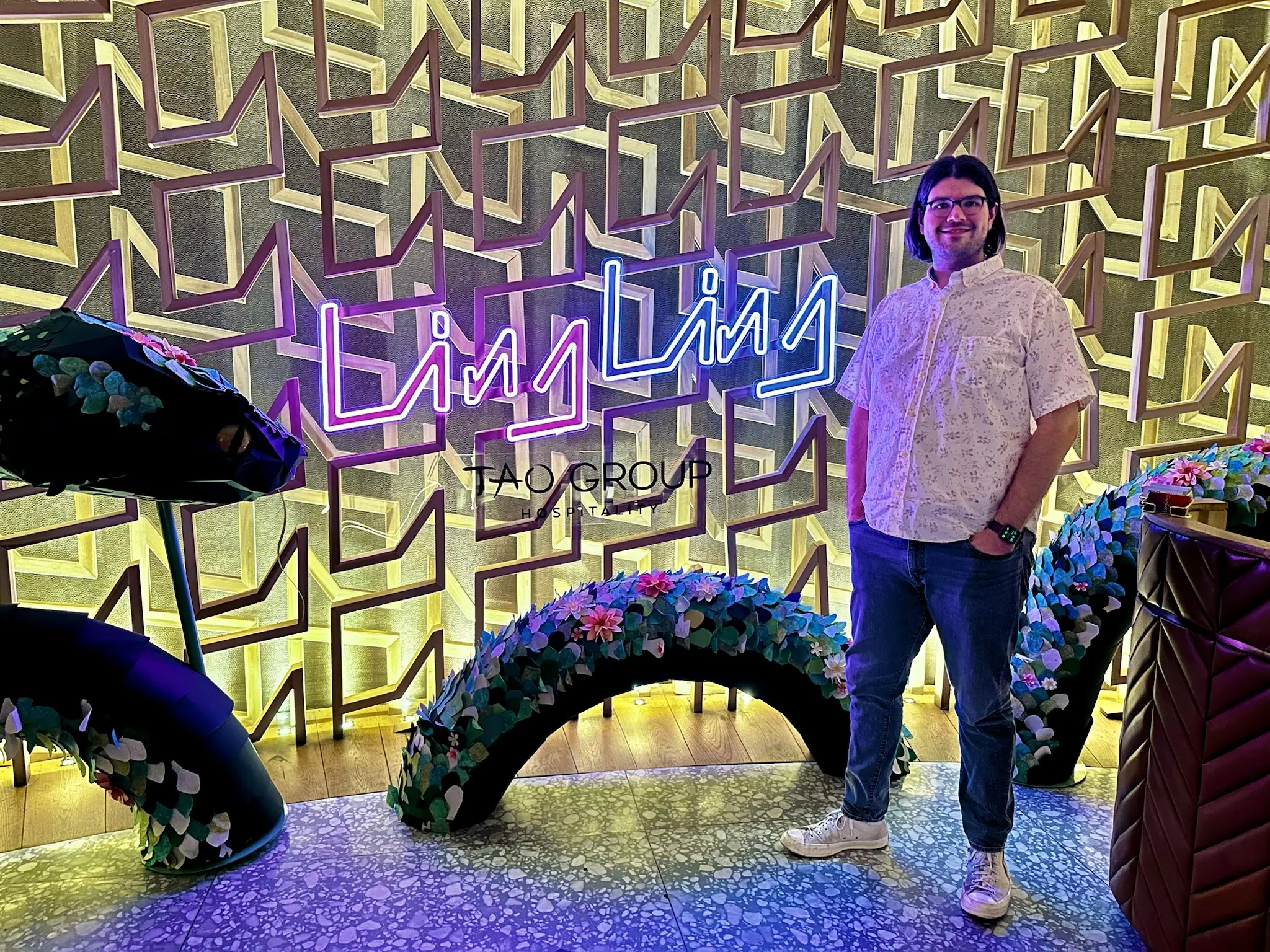 A dimly lit lobby with a patterned, textured wall with a pink and blue neon sign that says "Ling Ling" on the wall. Around the sign is a dragon made out of a topiary that looks like it's dissapearing into and out of the floor. To the right of the sign is a person (me!) with neck length black hair, blue glasses, and an Apple Watch, wearing a short sleeve white collared shirt, gray jeans, and white Converse high-tops.