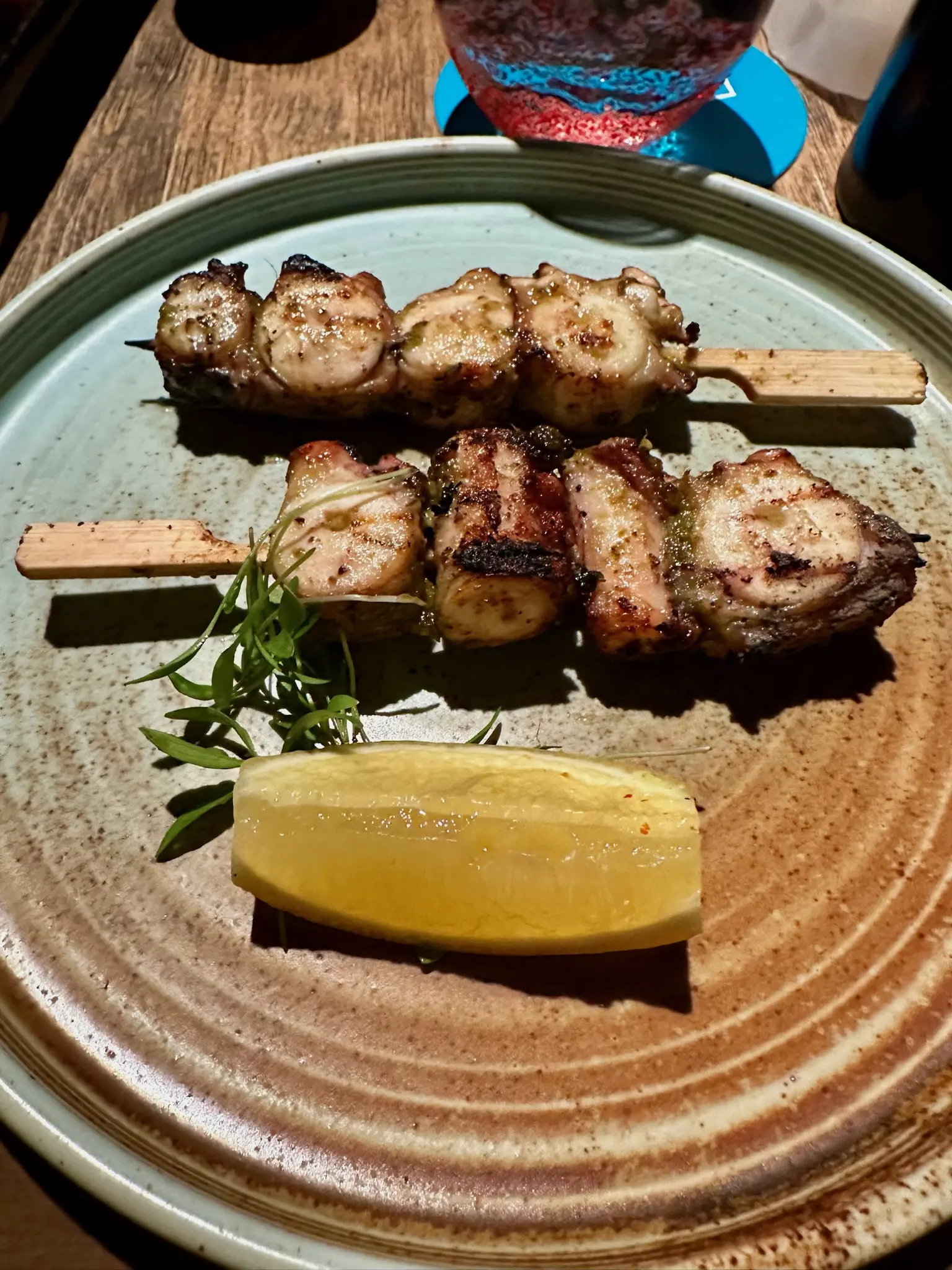 On a round brown ridged plate sit two skewers with four pieces of sliced octopus on each. In front of the skewers is a lemon slice and some sort of green garnish.