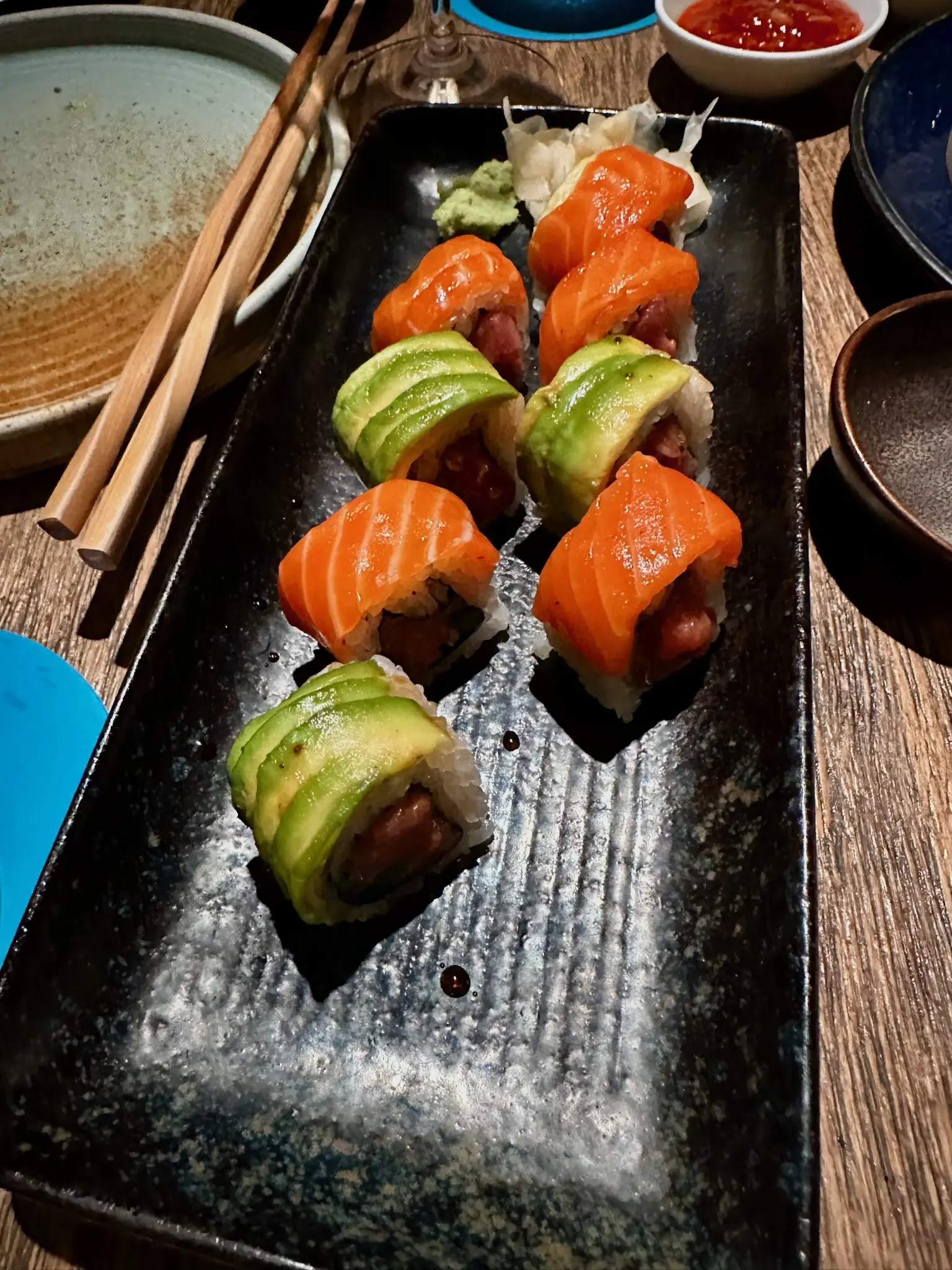 On a long rectangular black plate sit eight sushi roll slices. Three are topped with slices of avocado and five are topped with slices of salmon.