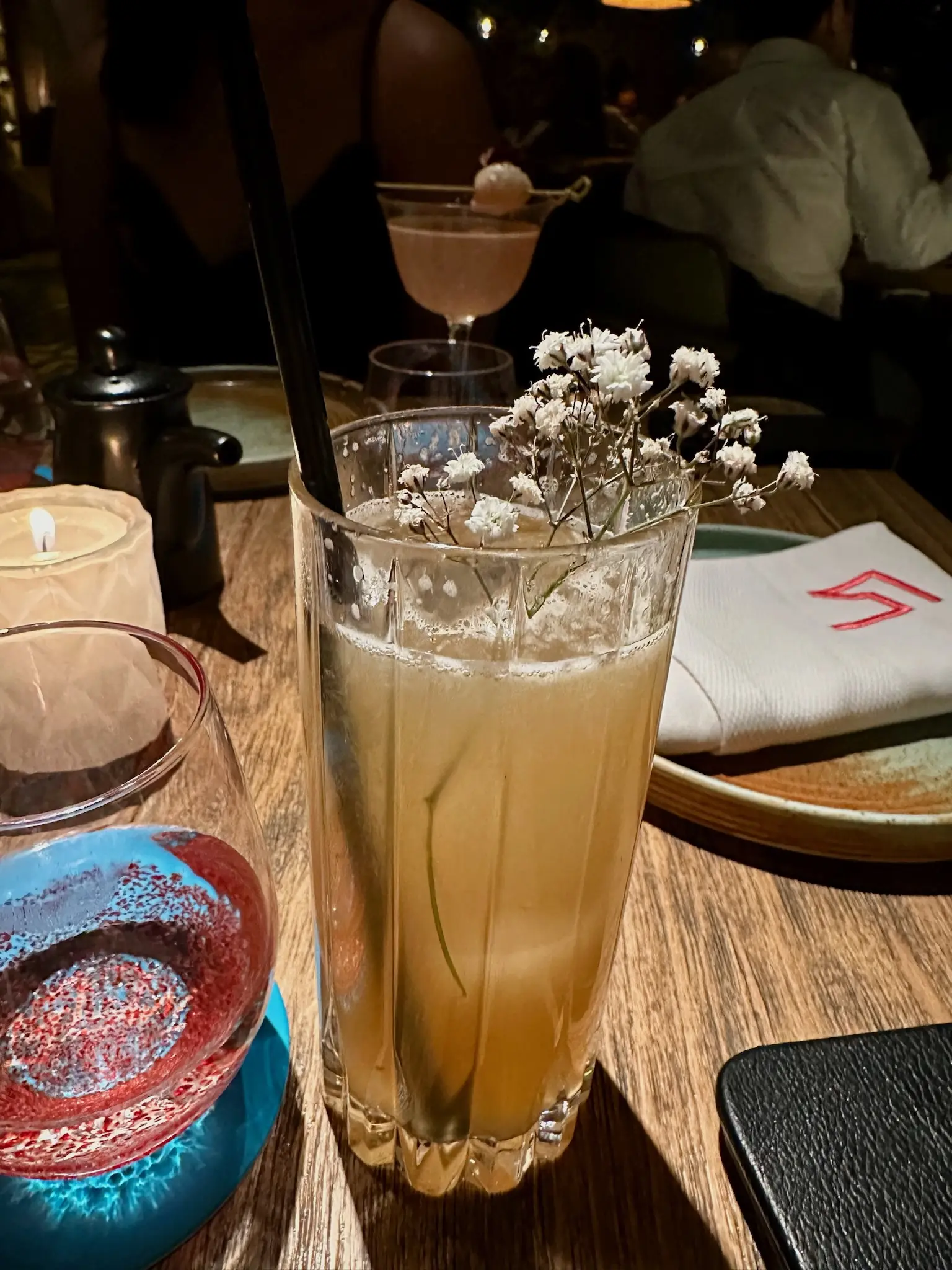 A Collins glass with a pale yellow drink and a black straw. In the drink is a small stem with seversl small white flowers on it.
