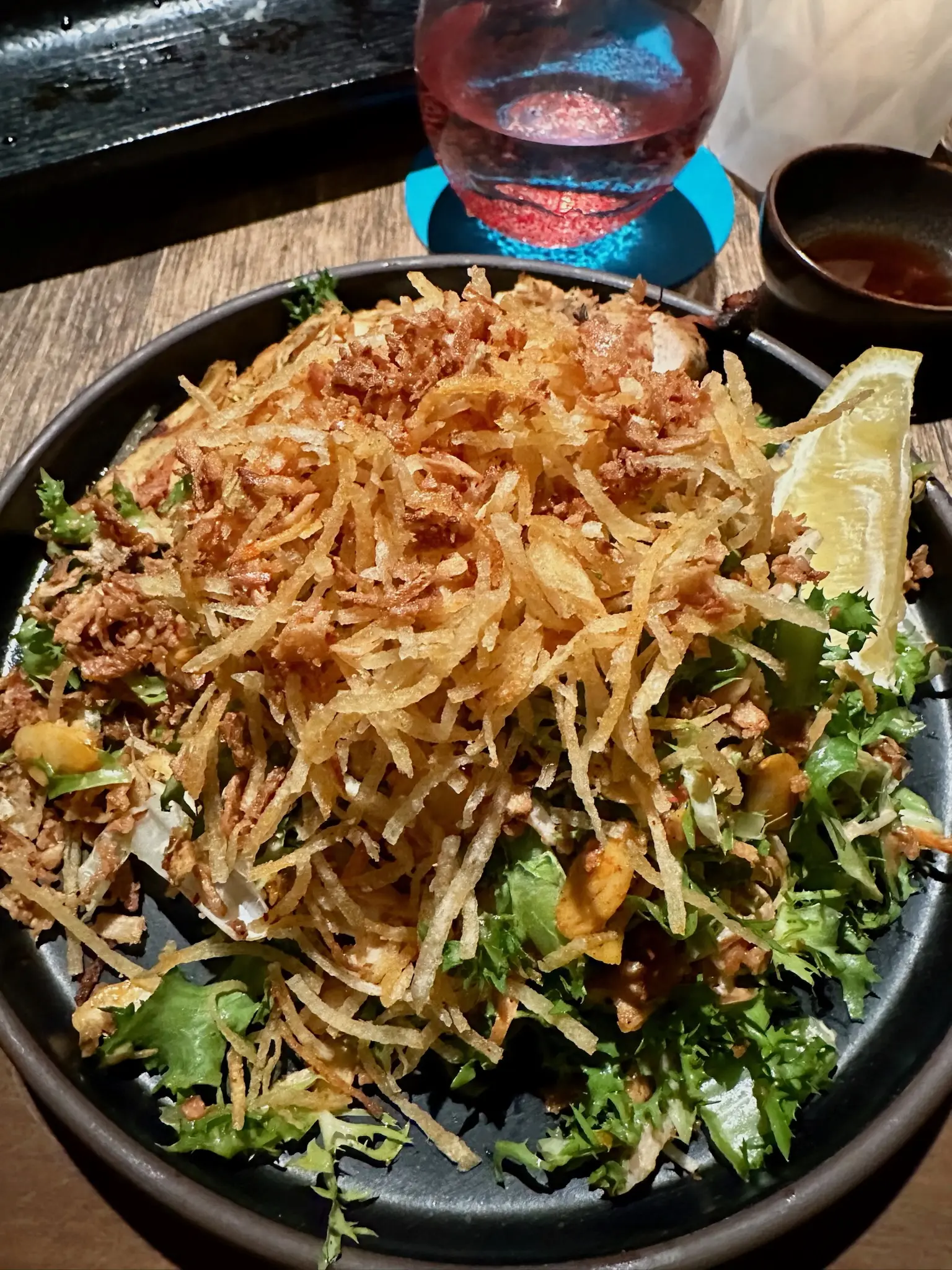 On a round black plate sits green lettuce, piled high with brown crispy strips of something or other and a visible lemon slice in the top right of the frame.