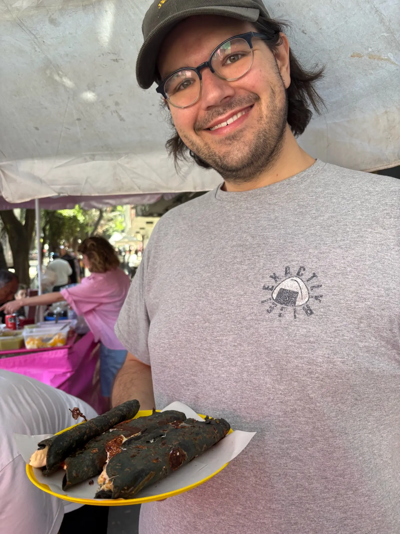 A person (me!) with neck length black hair and blue glasses, wearing a brown courderoy baseball cap with the words "yes, chef" in yellow printed on the front and gray tee-shirt with an onigiri in the upper left with the words "Exactly Rice!" around it, holds a plastic yellow plate with three blue corn quesadillas on it.