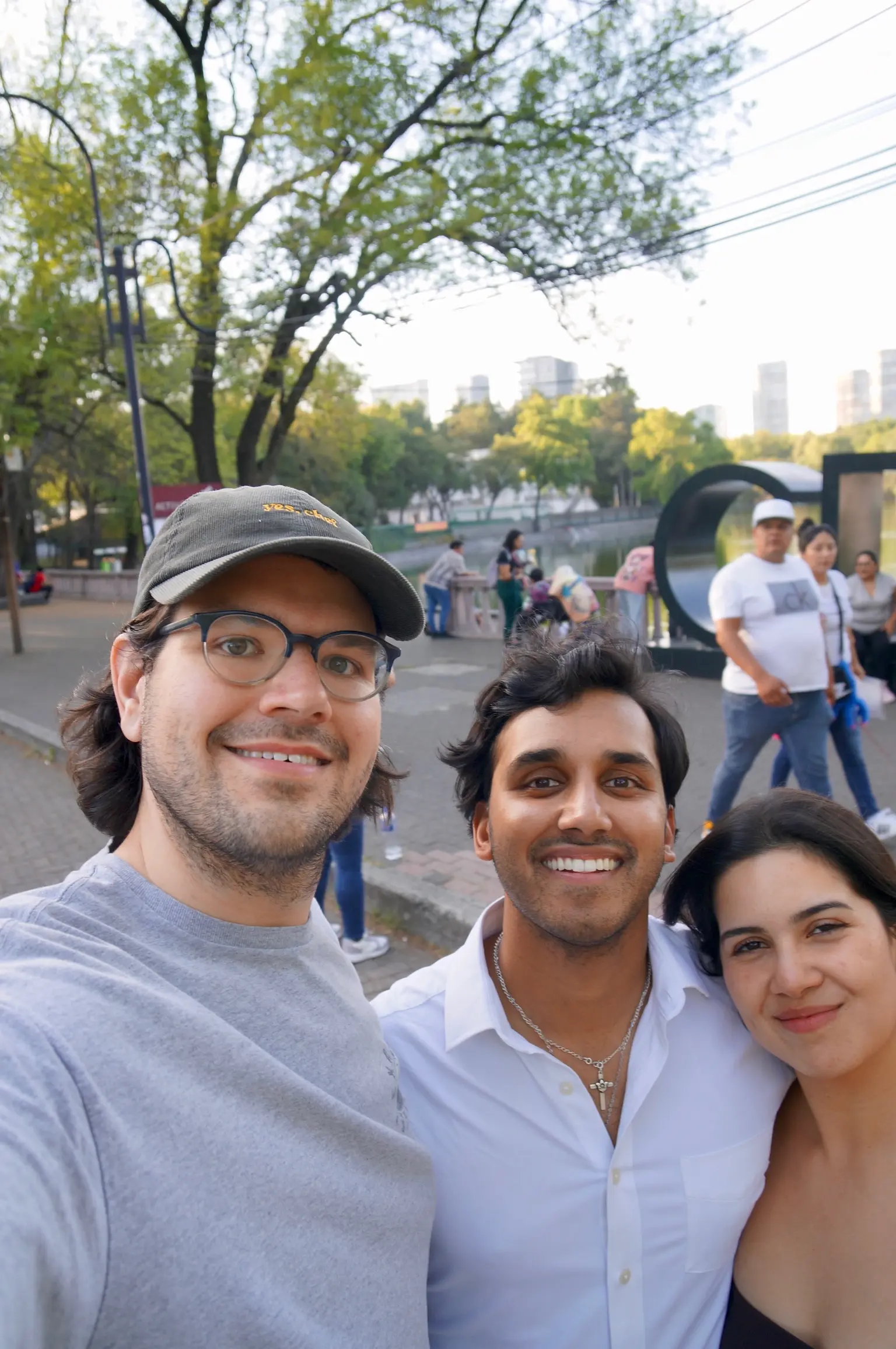 A person (me!) with neck length black hair and blue glasses, wearing a brown courderoy baseball cap with the words "yes, chef" in yellow printed on the front and gray tee-shirt stands next to a man in with shaggy black hair in a long sleeve white collared shirt with a cross necklace on and a woman with long black gair in a maroon dress. Behind them is a park, lake, and cityscape.