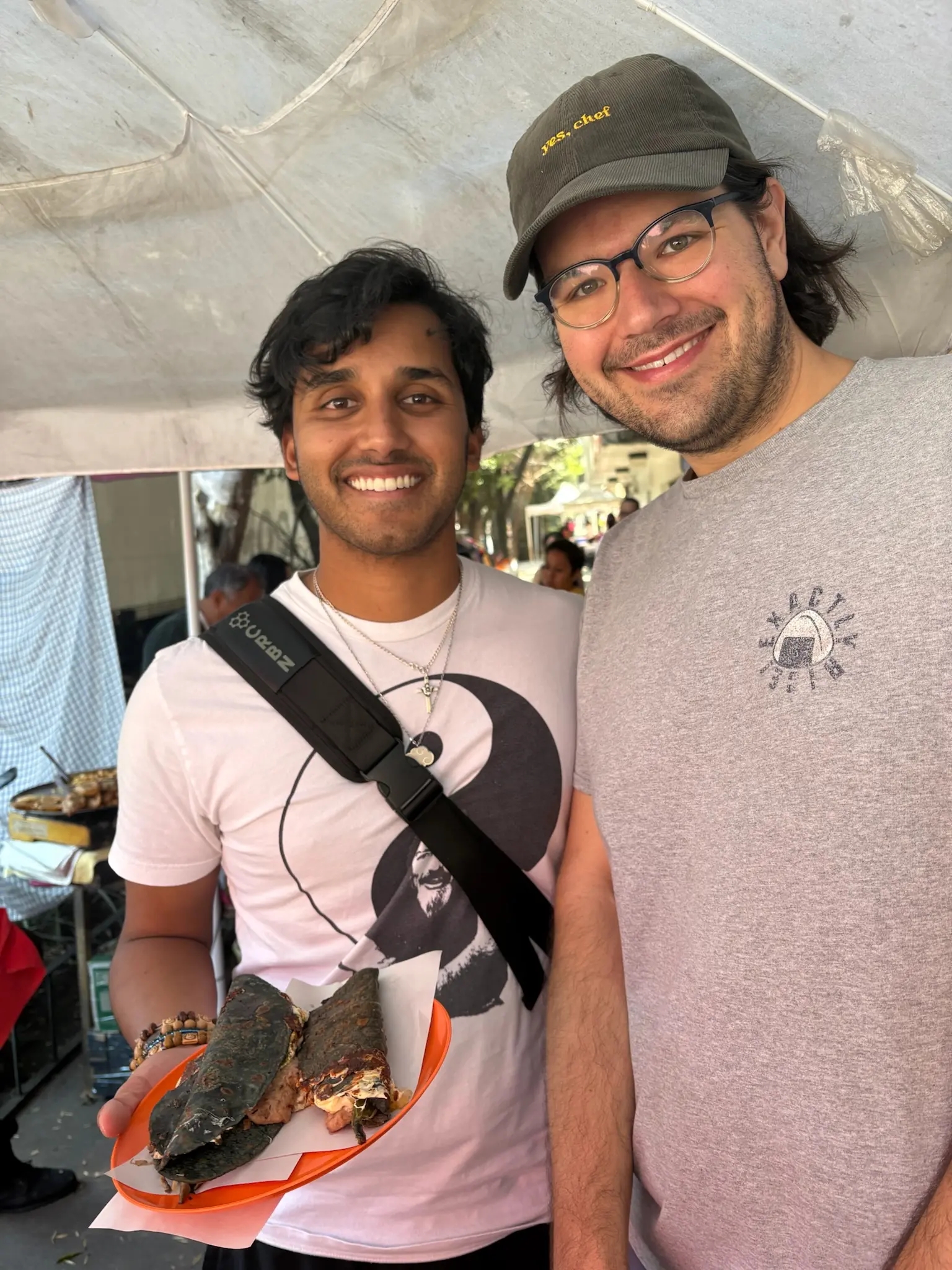 A person with shaggy black hair in a white shirt with a yin-yang on it holds a red plastic plate with two blue corn quesadillas on it. Next to him stands a person (me!) with neck length black hair and blue glasses, wearing a brown courderoy baseball cap with the words "yes, chef" in yellow printed on the front and gray tee-shirt with an onigiri in the upper left with the words "Exactly Rice!" around it.