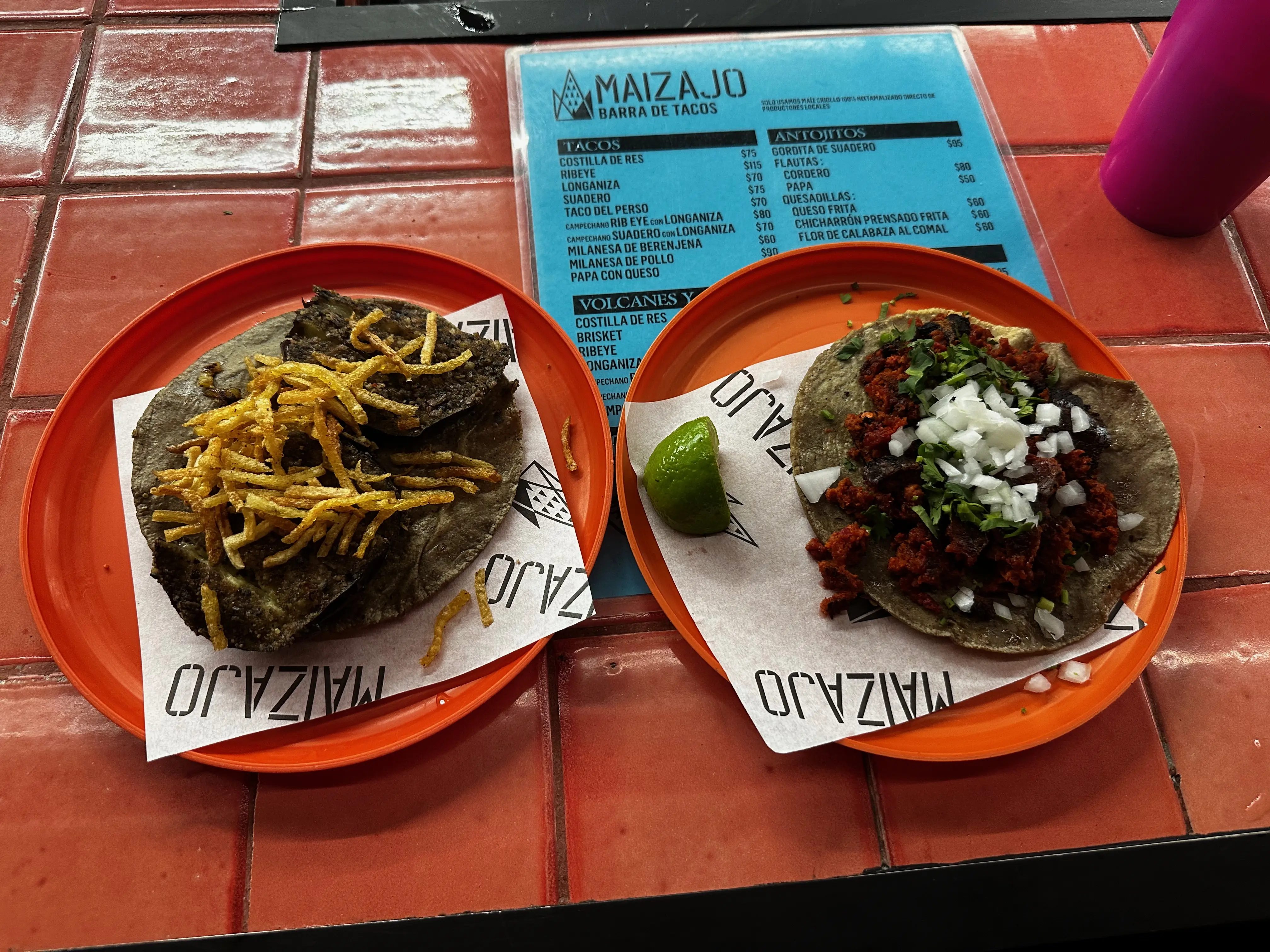On a counter of small, square, red tiles sit two plastic red plates. On top of the plates are a white paper with the word "MAIZAJO" printed on them. On the plate on the left sits a blue corn tortilla topped wih fried eggplant slices and thin slices of crispu fried potatoes. The plate on the right has a lime slice and a taco on a blue corn tortilla with red crumbled longaniza sausage, white onion, and cilantro on top. In the background is a pink cup filled with horchata and a blue menu sits under the plates.