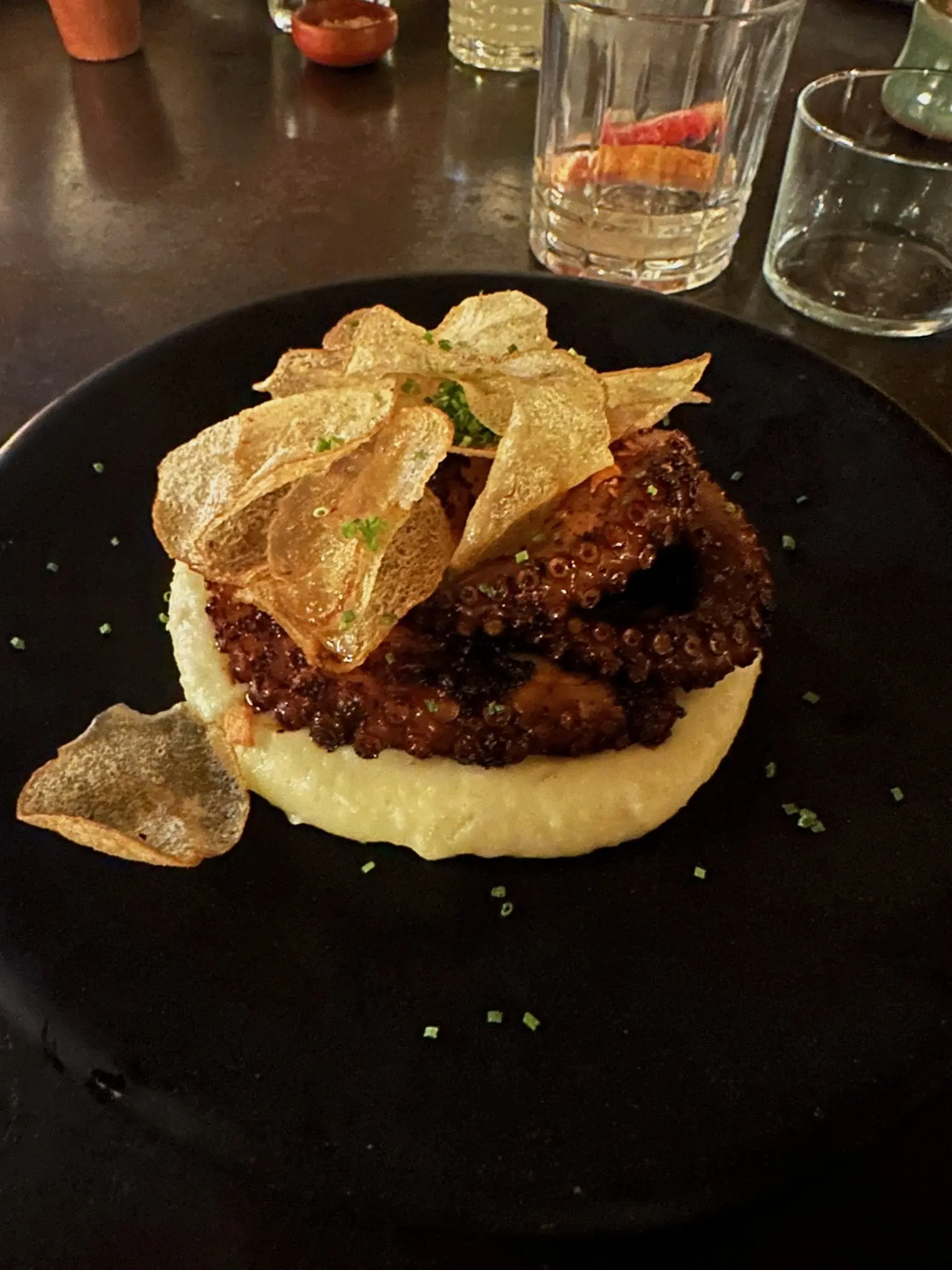 On a black plate sits a bed of yellow mashed motatoes. On top of the potatoes are grilled octopus tentacles. On top of the octopus are crispy fried potato slices.