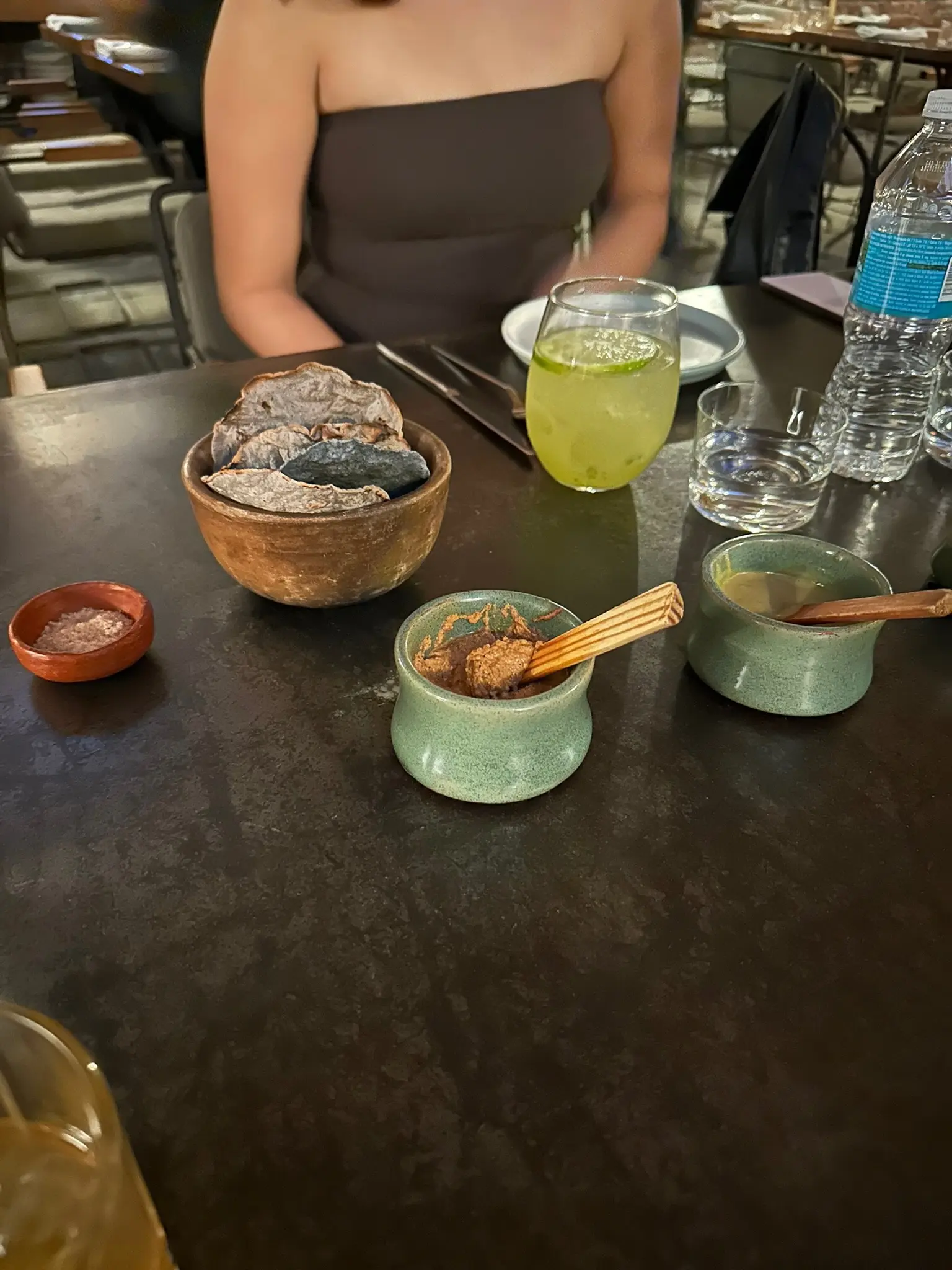 On a black table sits two identical round teal containers with salsa and wooden spoons in each. The one closest to the camera has a chunkier brown salsa in it and the one farther from the camera has a thinner, smoother green salsa. Behind them sit a wooden bowl of blue-corn tostadas and next to it is a small wooden bowl of salt.