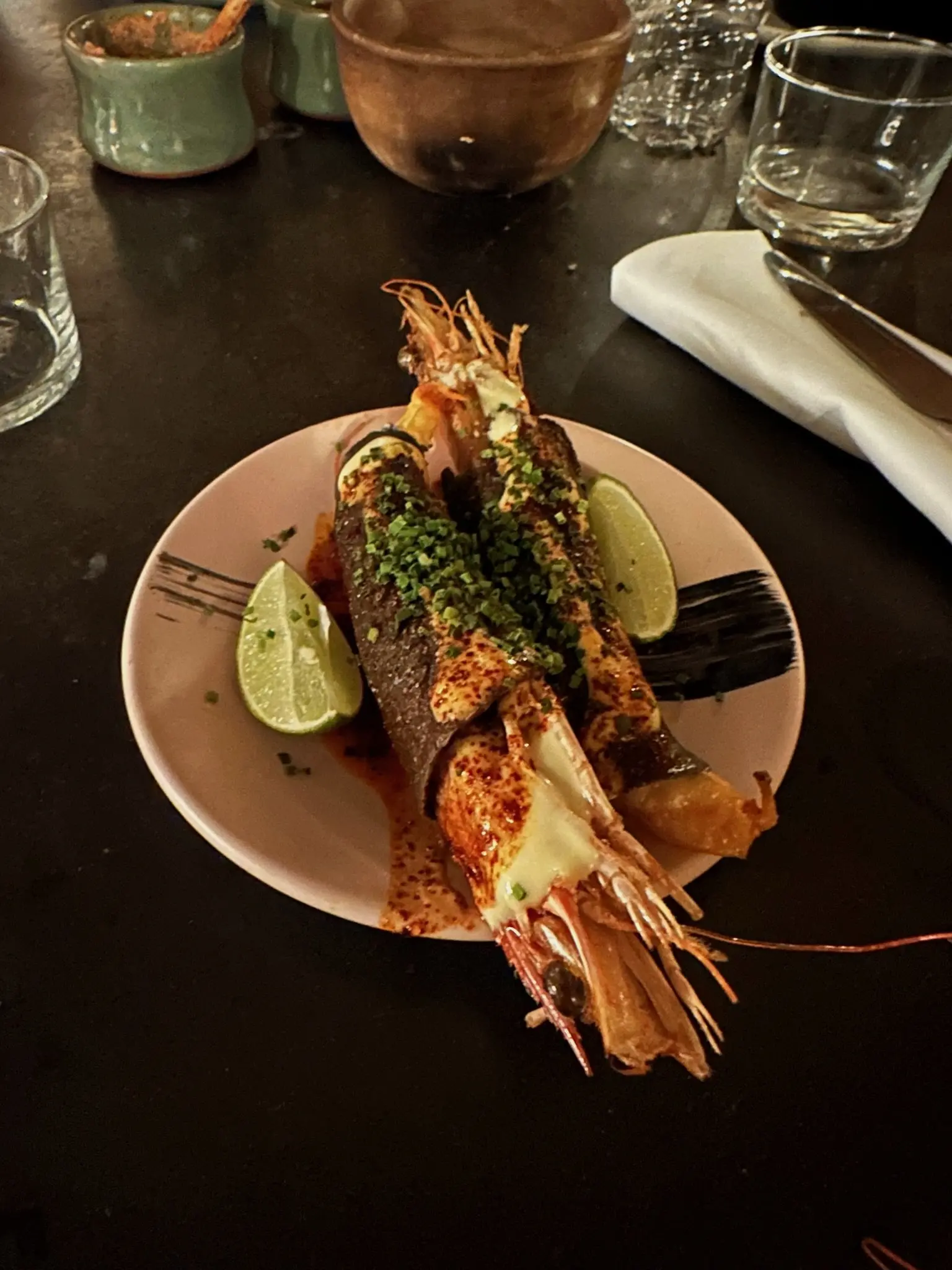 Two tacos on a white plate with lime slices on either side of them. The tortillas are blue corn and topped with chopped green chives. Inside the tortillas are cooked red shrimp with the heads and shells on.