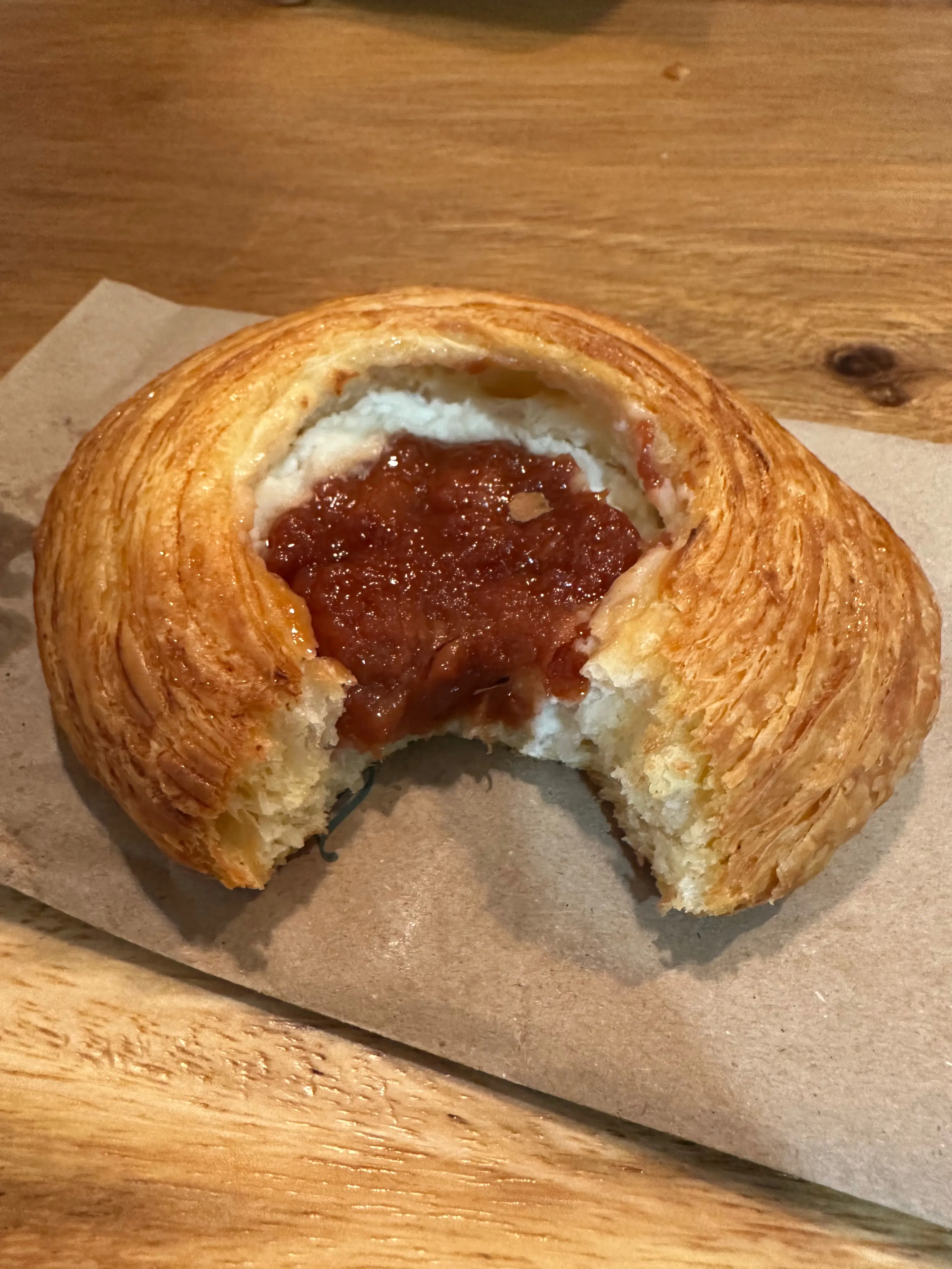 A circular, laminated pastry with cheese and guava in the middle sitting on a brown paper bag. A single bite is taken out of it.