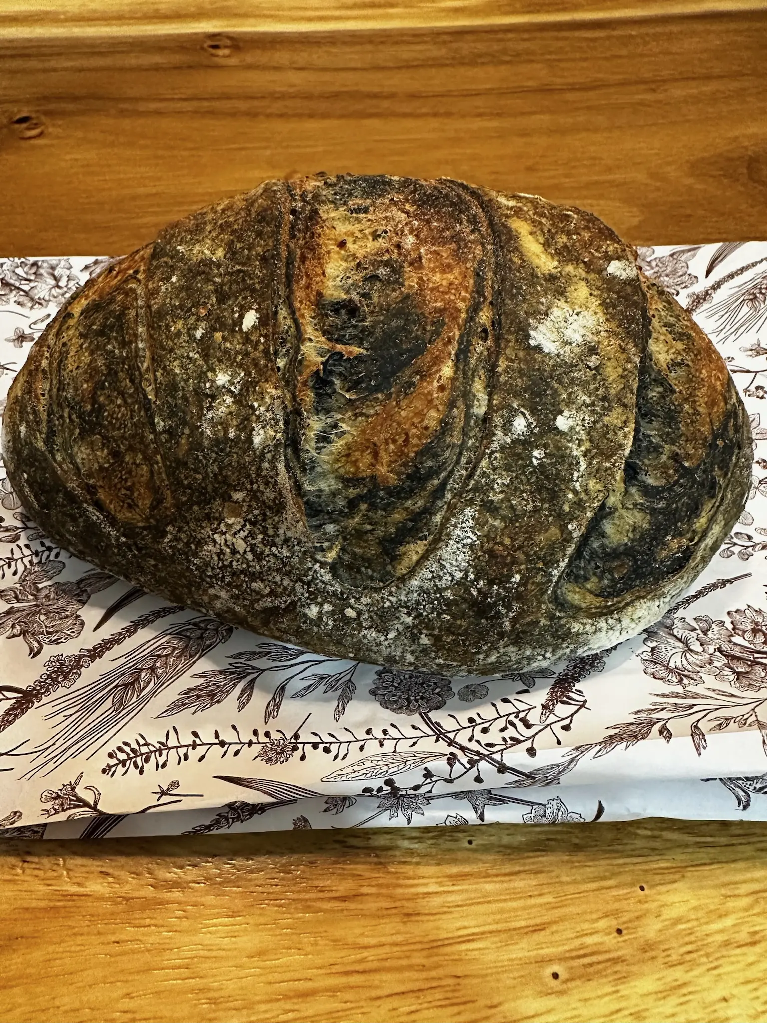 A loaf of sourdough bread which has been ripped in half, sitting on a white bag with black flowers and grains printed on it. The bread has a dark brown crust, with spekles of black swirls.