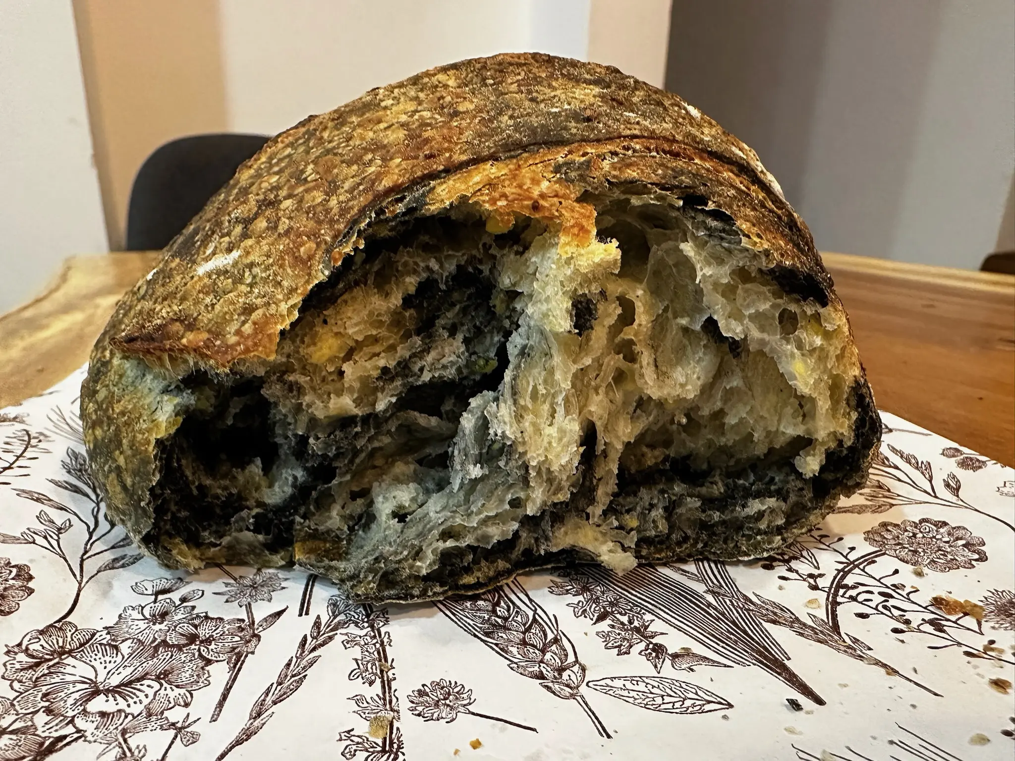 A loaf of sourdough bread which has been ripped in half, sitting on a white bag with black flowers and grains printed on it. The bread has a dark brown crust, with a light brown inside, swirled with black as well.