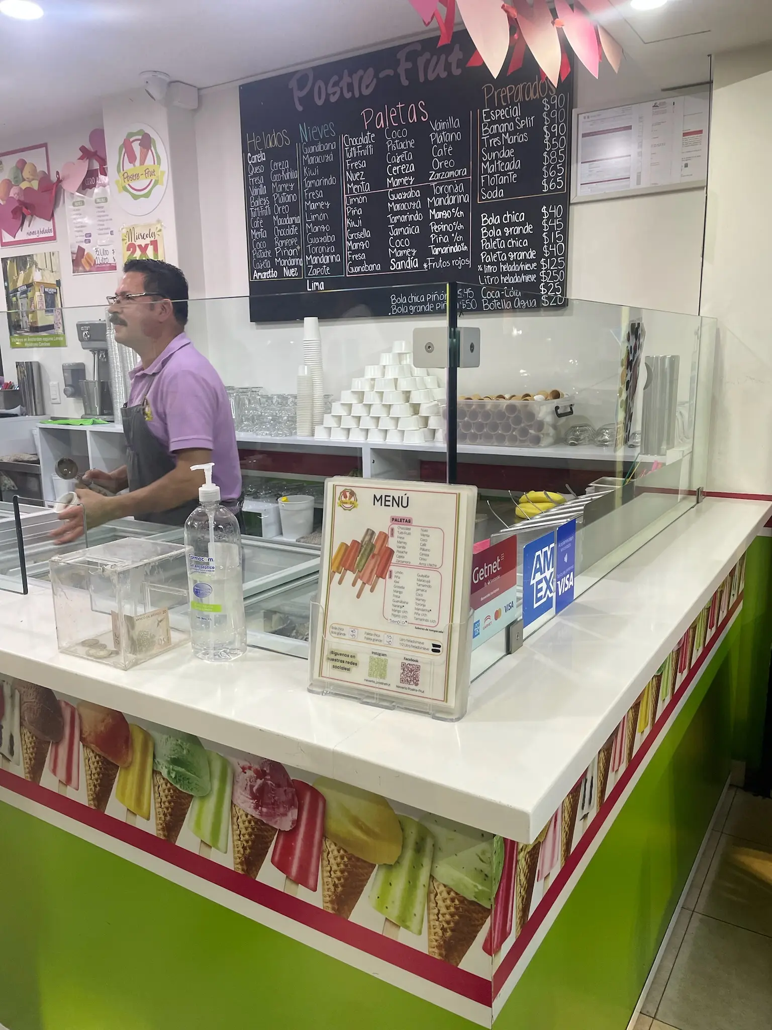 A lime green ice cream counter with a man with a mustache and a purple shirt behind it serving ice cream. Behind him is a black chalk board with hand-written ice cream flavors on it.