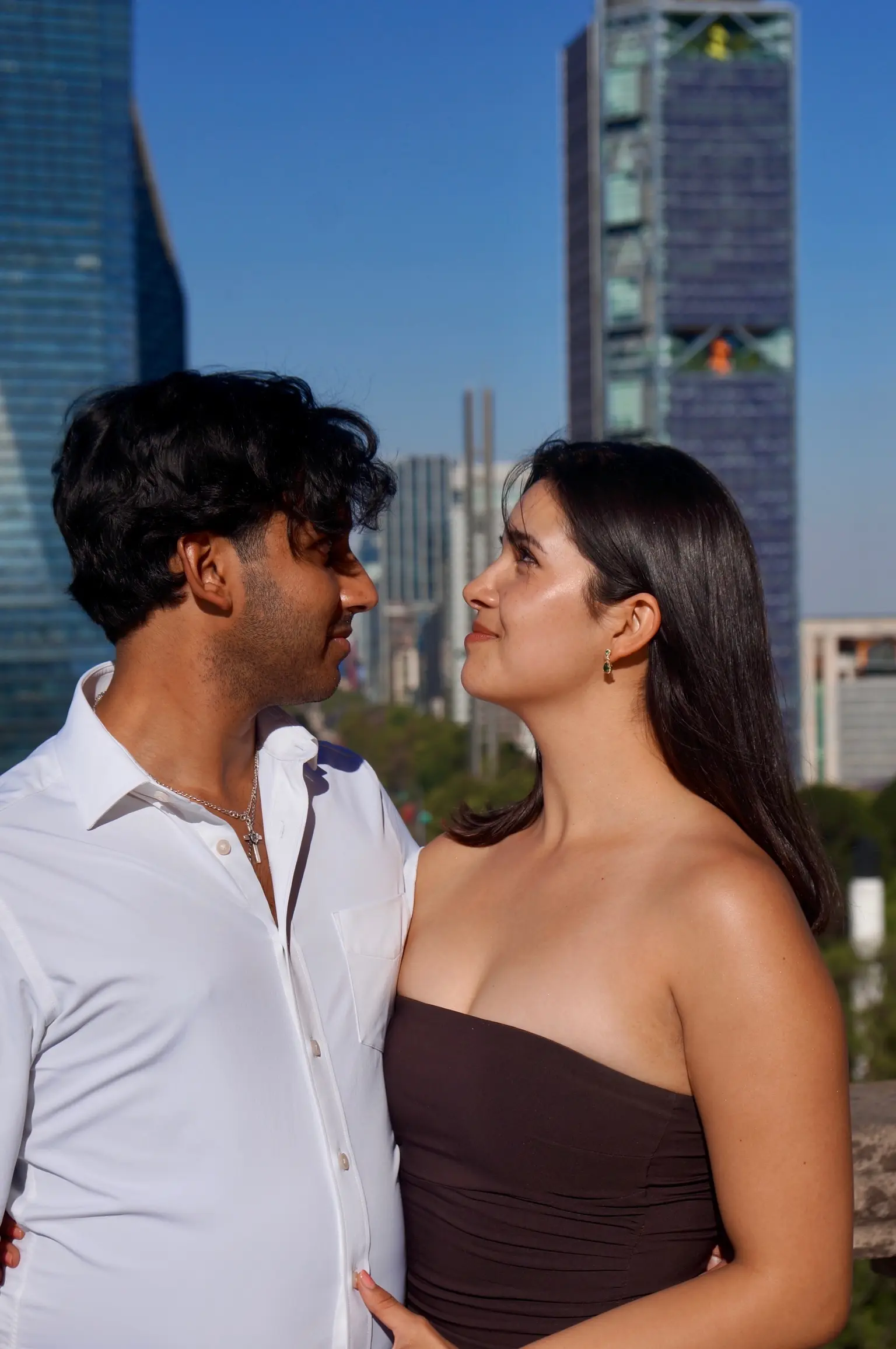 A man with short black hair in a white collared shirt faces a woman with long black hair in a maroon dress. Behind them are skyscrapers and a lush city avenue.